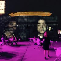 A photo of a theater with people waiting to enter and projected facial images on the side of the theater.