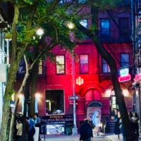 A small narrow street at night with a man walking down the middle of the street and a brigh red building at the far end.